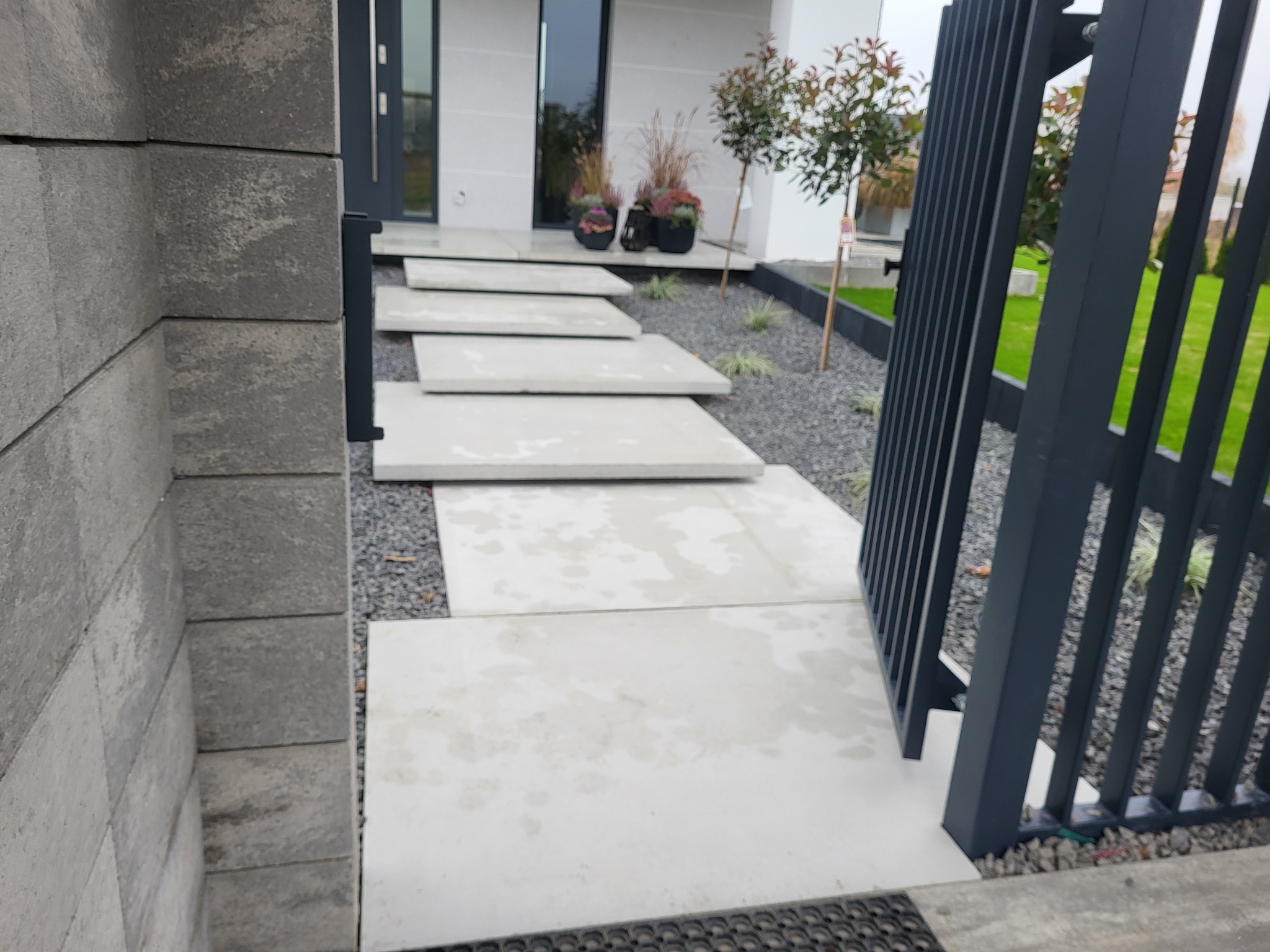 Nowoczesna ścieżka z płyt betonowych prowadząca do domu, wysypana drobnym kamieniem, z widocznym fragmentem ogrodzenia i elewacji budynku.