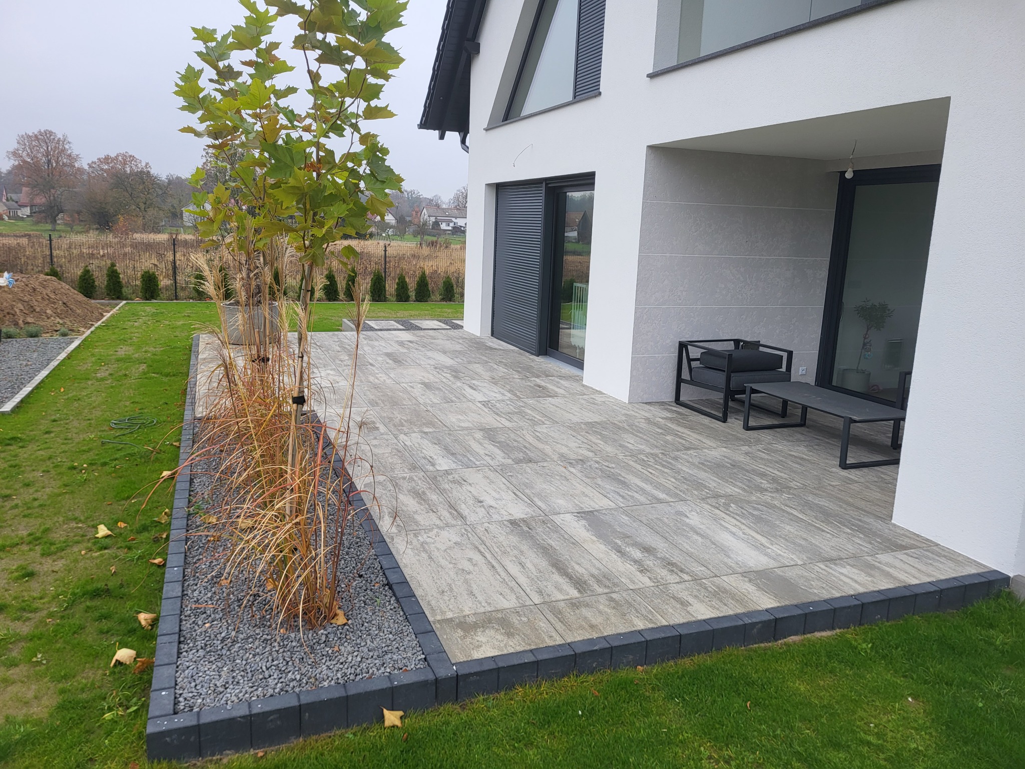 Nowoczesny taras z szarych płyt, obrzeża z ciemnej kostki. Minimalistyczne meble ogrodowe, trawa i drzewo w tle. Eleganckie wykończenie przestrzeni.