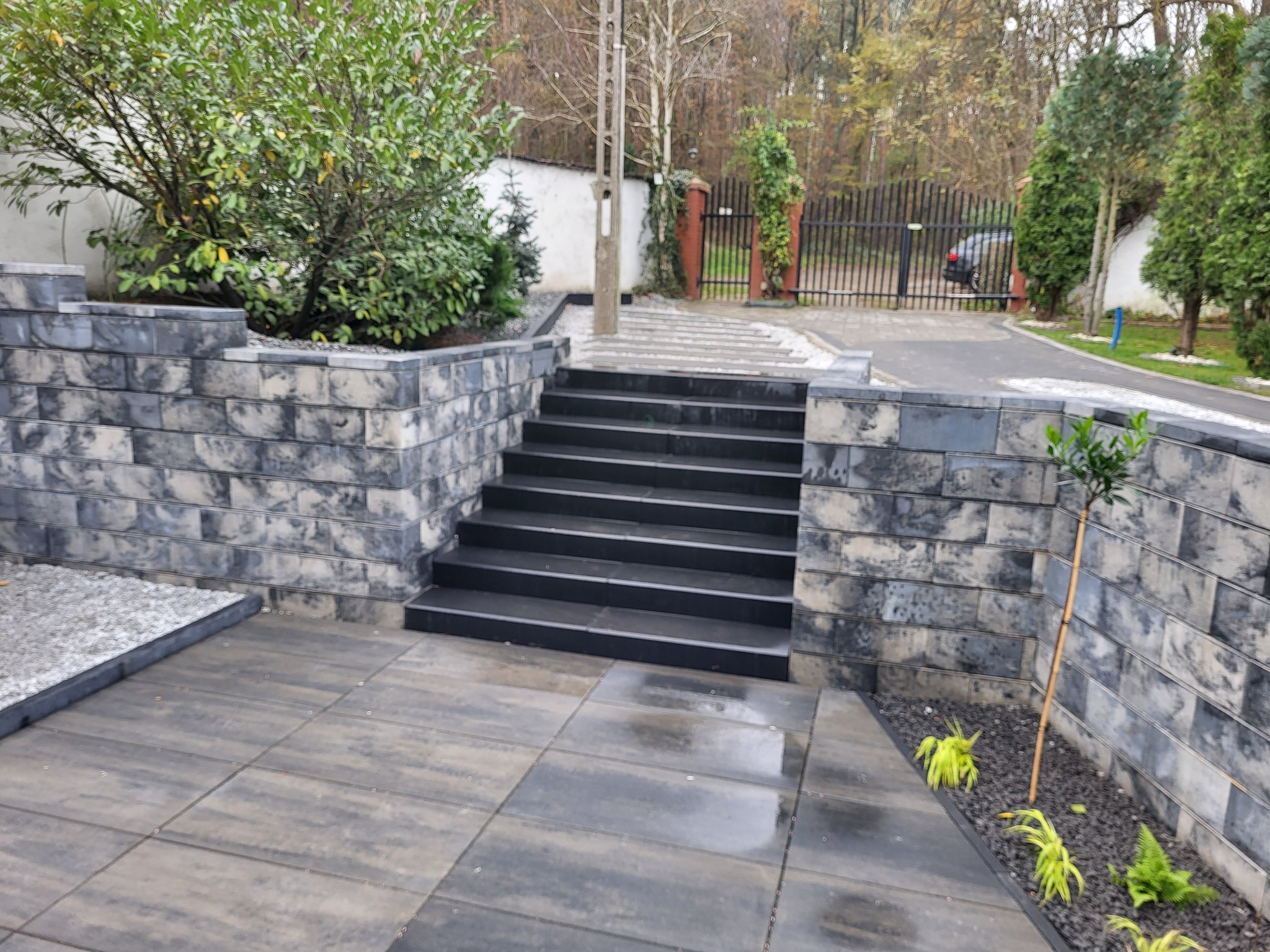 Schody z ciemnej kostki prowadzące do bramy, otoczone murem z szarej kostki. Uwagę przykuwają rośliny ozdobne wzdłuż muru i starannie ułożona nawierzchnia.