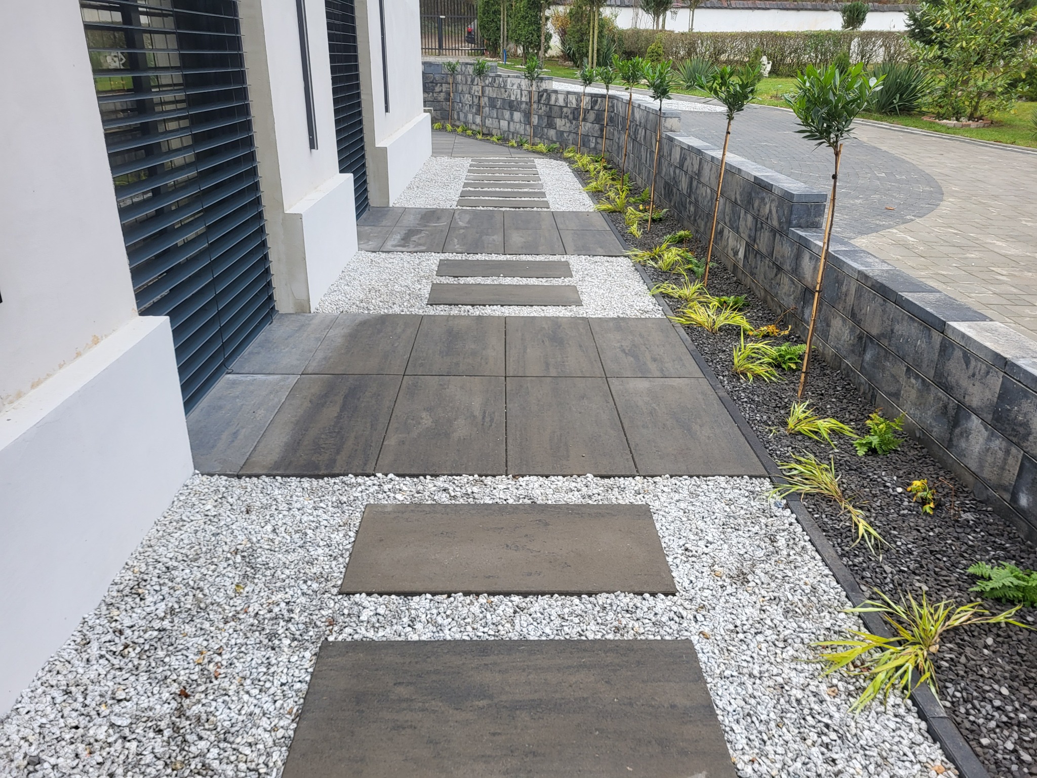 Nowoczesna ścieżka z szarych płyt i białego grysu prowadząca wzdłuż budynku z żaluzjami, obok murku z nasadzeniami. Estetyczne wykończenie terenu.