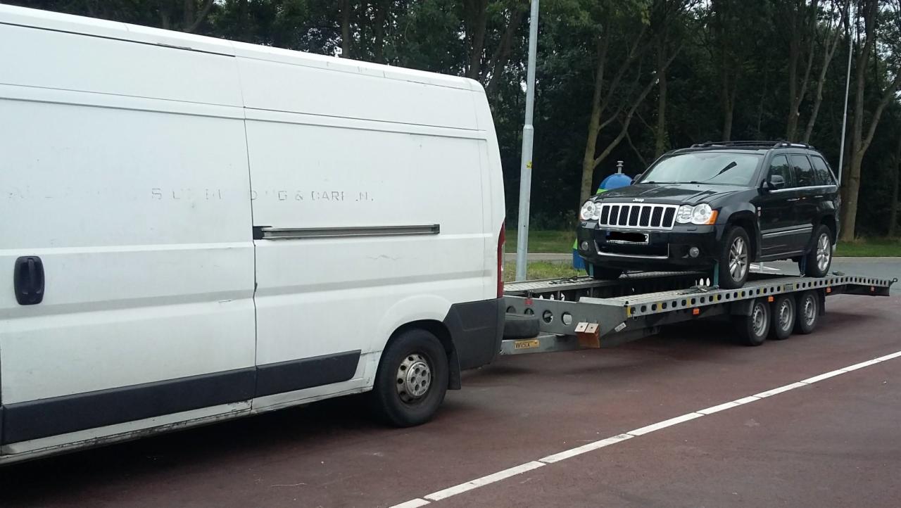 Czarny Jeep Grand Cherokee przewożony na lawecie z trzema osiami, ciągniętej przez biały bus z ledwo widocznym napisem 'SUPRI D'G & GARI N.'