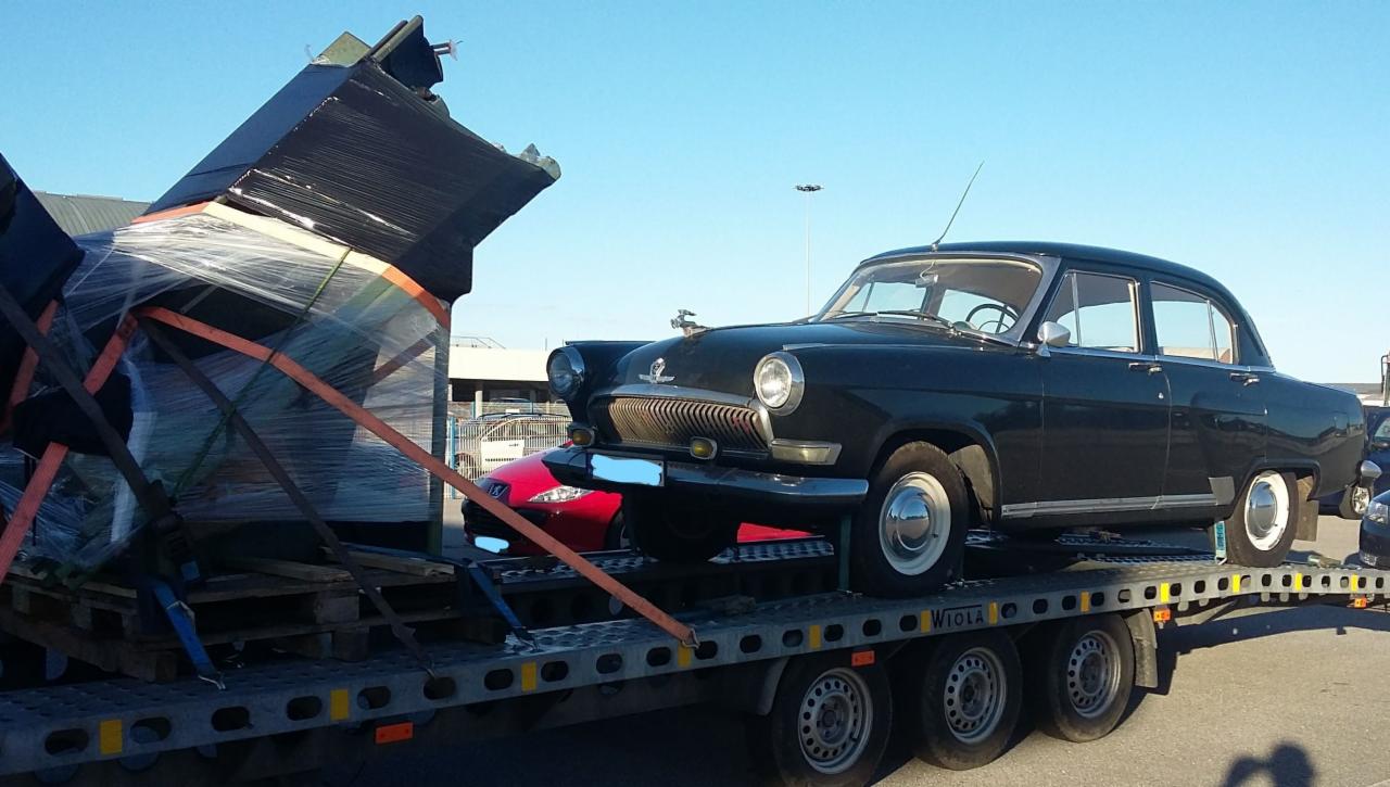 Czarna Wołga GAZ-21 na lawecie, zabezpieczona pasami, obok zapakowane meble na paletach, również zabezpieczone folią stretch.