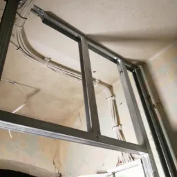 Instalacja elektryczna w trakcie remontu: widoczne przewody przymocowane do sufitu i metalowa konstrukcja podwieszanego sufitu, stara ściana z widocznymi pęknięciami.