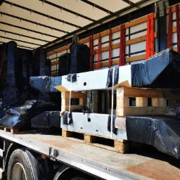 Załadunek konstrukcji stalowych, zabezpieczonych czarną folią, na platformę transportową ciężarówki, widoczne drewniane palety i elementy mocujące.