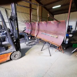 Spawanie segmentu konstrukcji stalowej w warsztacie, widoczny wózek widłowy i spawarka, surowa powierzchnia metalu z widocznymi spawami.