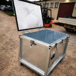 Otwarty, ocynkowany kontener stalowy z widocznymi zatrzaskami, umieszczony na żwirowym podłożu, z budynkiem i wózkiem widłowym w tle.