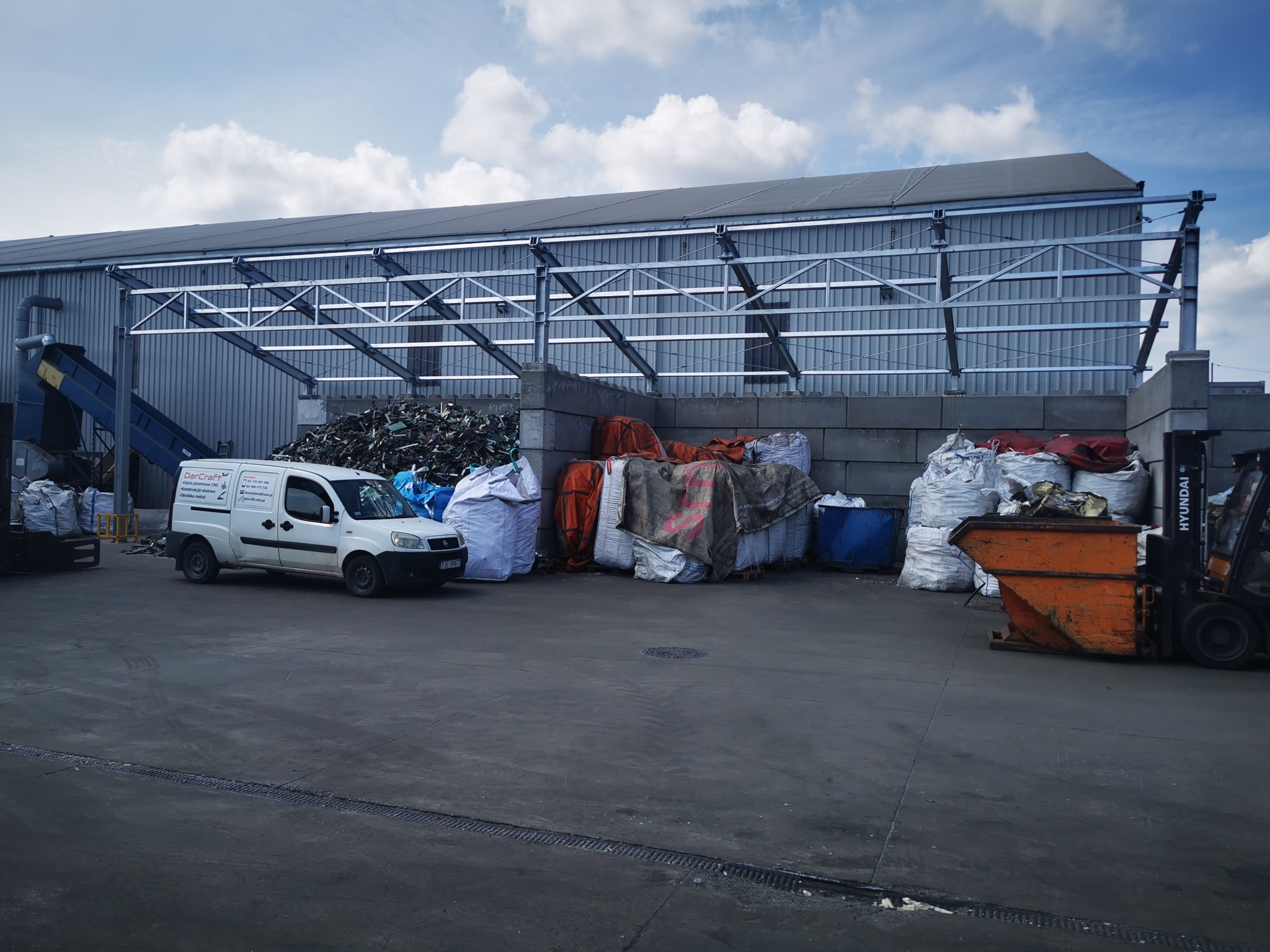 Zewnętrzny widok hali magazynowej o konstrukcji stalowej z zaparkowanym białym vanem z logo firmy, workami z odpadami i pomarańczowym kontenerem na odpady obsługiwanym przez wózek widłowy.