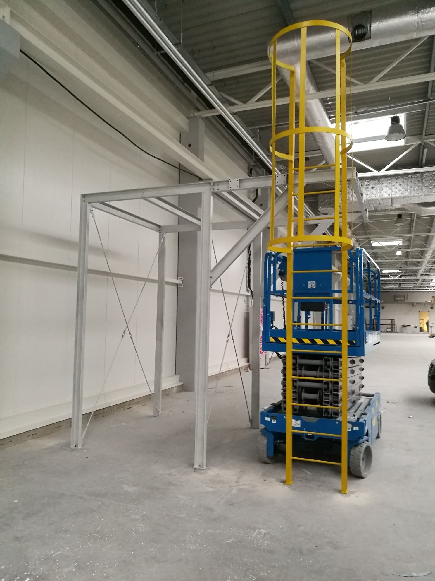 Wnętrze hali w trakcie budowy, widoczna stalowa konstrukcja szkieletowa i podnośnik nożycowy z żółtą klatką bezpieczeństwa.
