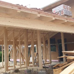 Surowa konstrukcja domu szkieletowego z widocznymi drewnianymi belkami, częściowo obłożona płytami, z zapasem dachówek na dachu i stertą materiałów budowlanych obok.