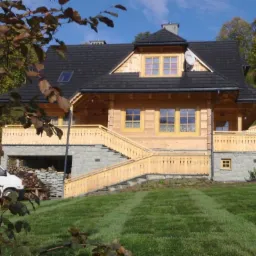 Nowoczesny, drewniany dom z kamienną podmurówką, z zadbanym trawnikiem z widocznymi pasami po koszeniu, częściowo przysłonięty gałęziami drzewa