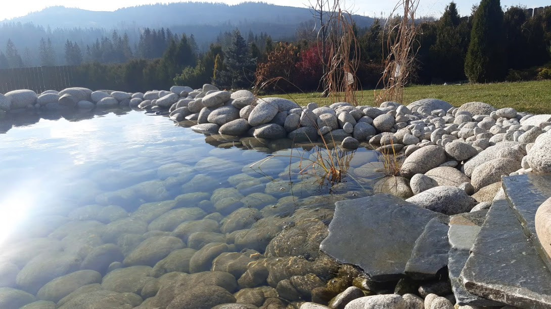 Oczko wodne wyłożone otoczakami i płytami skalnymi, z widokiem na zalesione wzgórza w tle, w słoneczny dzień.