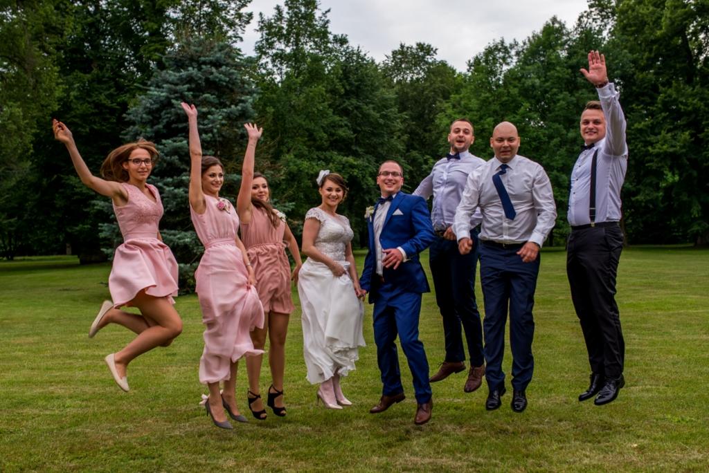 Uśmiechnięta para młoda i goście weselni w strojach w odcieniach różu i granatu, skaczący radośnie na tle zielonego trawnika w plenerze.