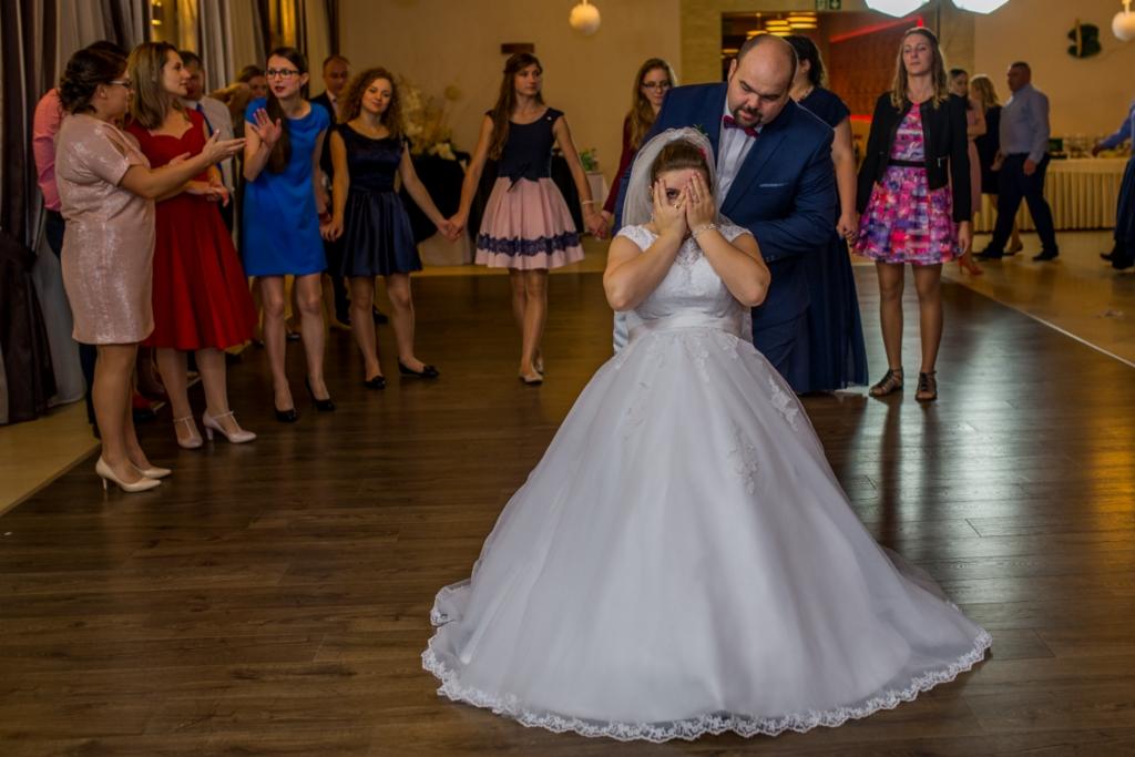 Panna młoda w białej sukni klęczy na parkiecie, zasłaniając twarz dłońmi podczas oczepin, w tle goście weselni i pan młody w granatowym garniturze.