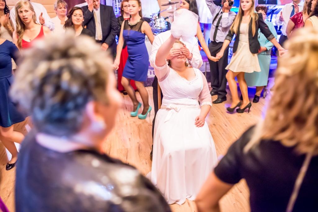 Panna młoda siedzi na krześle z zasłoniętymi oczami, otoczona tańczącymi gośćmi weselnymi podczas tradycyjnego oczepin na parkiecie z drewnianych paneli.