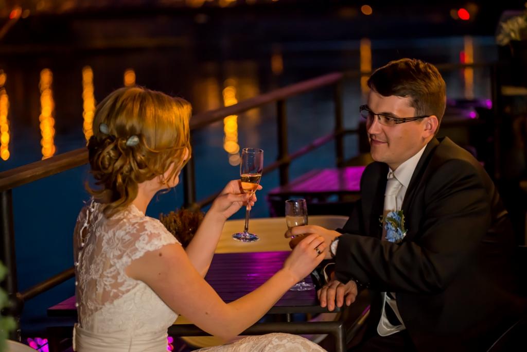 Para młoda wznosi toast szampanem na tle nocnego miasta, widoczne odbicia świateł w wodzie, ona w koronkowej sukni, on w czarnym garniturze z butonierką.