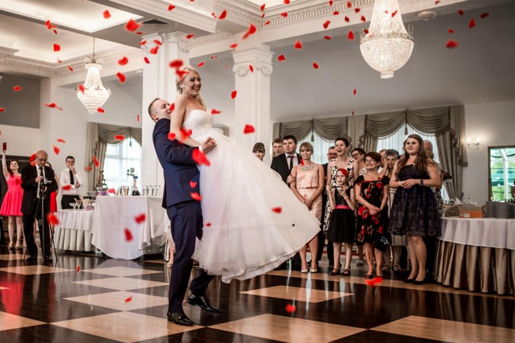 Pan młody podnosi uśmiechniętą pannę młodą w białej sukni podczas pierwszego tańca, w tle goście i opadające płatki róż.