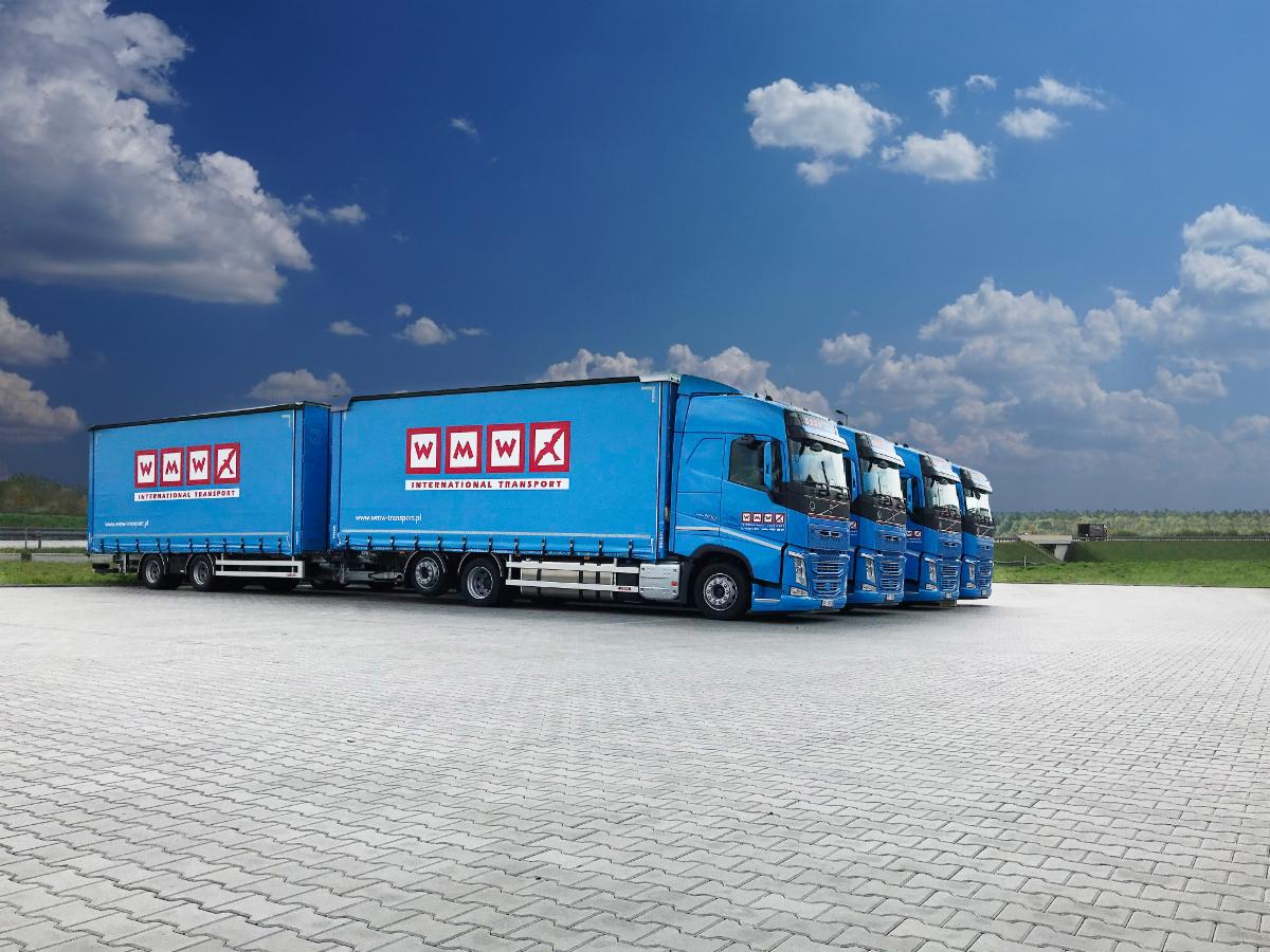 Flota czterech identycznych, błękitnych ciężarówek z naczepami, ustawionych w rzędzie na betonowym placu, z logo firmy transportowej na naczepach i kabinach, widok z boku, błękitne niebo z chmurami...