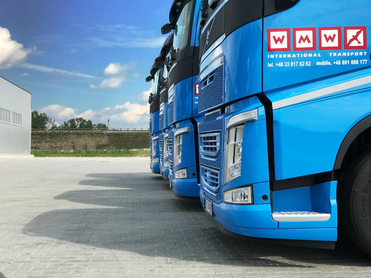 Szereg błękitnych ciężarówek z logo firmy transportowej na tle błękitnego nieba z chmurami, stojących na szarym brukowanym placu.