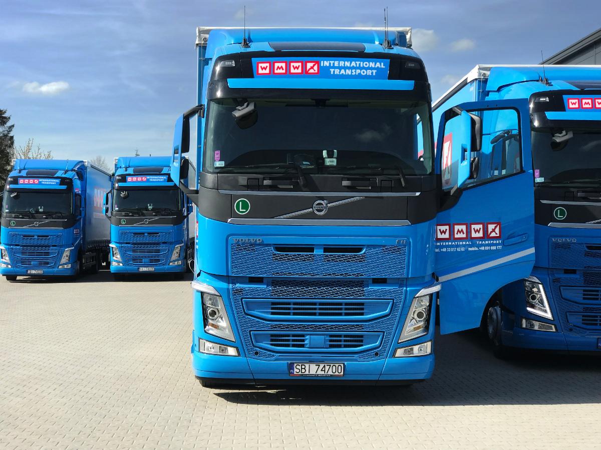 Flota niebieskich ciężarówek Volvo z logo firmy transportowej WMW International Transport, zaparkowana na placu z kostki brukowej w słoneczny dzień.