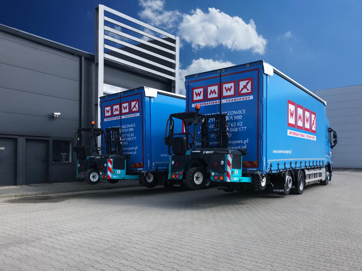 Dwa niebieskie samochody ciężarowe z logo firmy WMW International Transport zaparkowane przed magazynem, z widocznymi wózkami widłowymi Moffett zamontowanymi z tyłu.