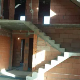 Wnętrze w trakcie budowy: surowe ściany z cegły, betonowe schody bez wykończenia, widoczne przewody elektryczne wystające ze ściany, okna bez szyb, światło dzienne wpadające do pomieszczenia.