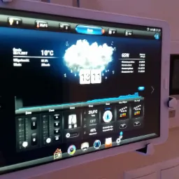 Tablet Samsung zamontowany na ścianie, wyświetlający interfejs systemu inteligentnego domu z informacjami o temperaturze, wilgotności, zużyciu energii i kontroli urządzeń, obok widoczny panel...