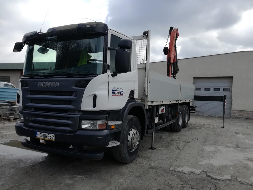 Ciężarówka Scania P380 z dźwigiem hydraulicznym na skrzyni ładunkowej, zaparkowana na betonowym placu przed budynkiem z bramą garażową.