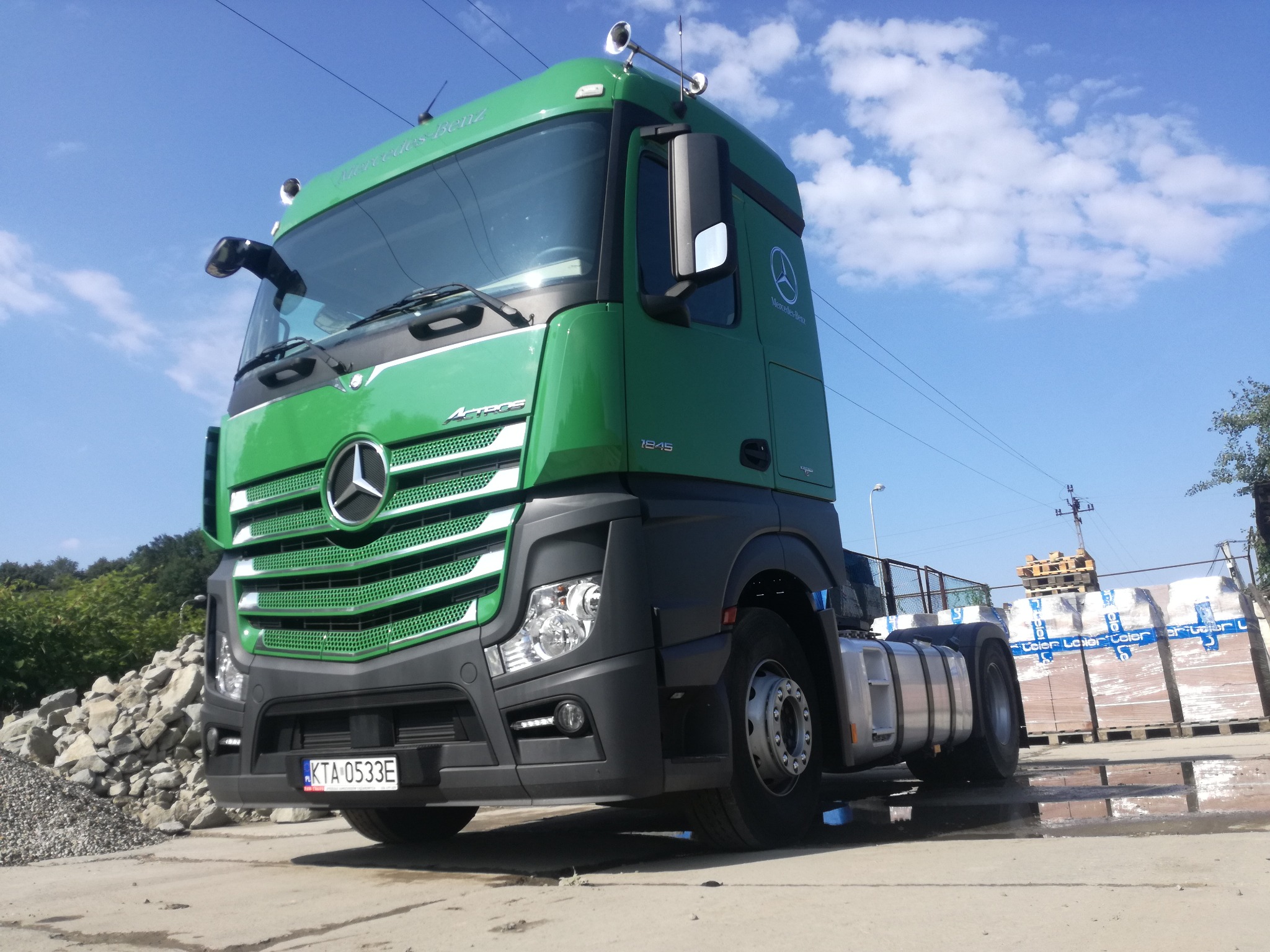 Zielony ciągnik siodłowy Mercedes-Benz Actros 1845 z widoczną tablicą rejestracyjną KTA 0533E, zaparkowany na betonowym placu z paletami towaru w tle.