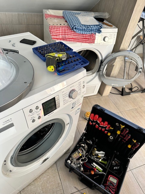 Rozłożona pralka z otwartymi drzwiczkami, obok skrzynka narzędziowa i wkrętarka. Widoczne ślady użytkowania na obudowie. W tle suszarka z tekstyliami.