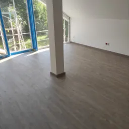 Jasne, przestronne wnętrze po remoncie z panelami podłogowymi w odcieniach szarości i białymi ścianami. Widoczne okna z niebieskimi ramami i filar w centrum pokoju.