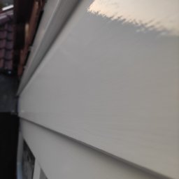 LUX&ART WNĘTRZA ŁUKASZ WĘGLOWSKI - Świeżo pomalowana, biała elewacja domu z widoczną fakturą paneli bocznych, uchwycona pod kątem z bliska. Widoczny fragment dachu z dachówką.