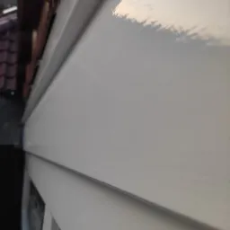 Świeżo pomalowana, biała elewacja domu z widoczną fakturą paneli bocznych, uchwycona pod kątem z bliska. Widoczny fragment dachu z dachówką.