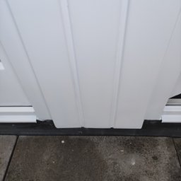 LUX&ART WNĘTRZA ŁUKASZ WĘGLOWSKI - Fragment białej, świeżo pomalowanej elewacji z widoczną listwą przypodłogową i szarą kostką brukową u dołu kadru. Ujęcie z dołu.