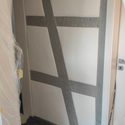 LUX&ART WNĘTRZA ŁUKASZ WĘGLOWSKI - Ściana z drewnianą konstrukcją pomalowana na biało i szaro, z widocznymi narzędziami malarskimi na podłodze, zabezpieczona folią malarską.