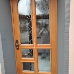 LUX&ART WNĘTRZA ŁUKASZ WĘGLOWSKI - Odnowione drewniane drzwi z szybami o zdobieniach, po renowacji. Widoczne ślady farby i odbicie osoby robiącej zdjęcie. Ściana pomalowana na szaro.