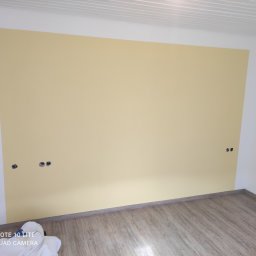 LUX&ART WNĘTRZA ŁUKASZ WĘGLOWSKI - Pomalowana na jasnożółto ściana w pokoju z panelami podłogowymi i gniazdkami elektrycznymi bez osprzętu. Widoczne zabezpieczenia podłogi.