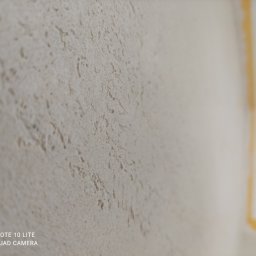 LUX&ART WNĘTRZA ŁUKASZ WĘGLOWSKI - Szpachlowana ściana o nieregularnej fakturze w odcieniach beżu, z widocznym fragmentem żółtej taśmy malarskiej przy suficie. Tekstura ściany jest wyraźnie widoczna.