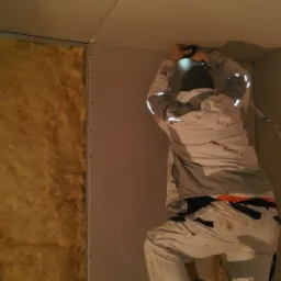 Pracownik w Lędyczku montuje płytę gipsowo-kartonową na ścianie z izolacją, używając wkrętarki i oświetlenia roboczego.