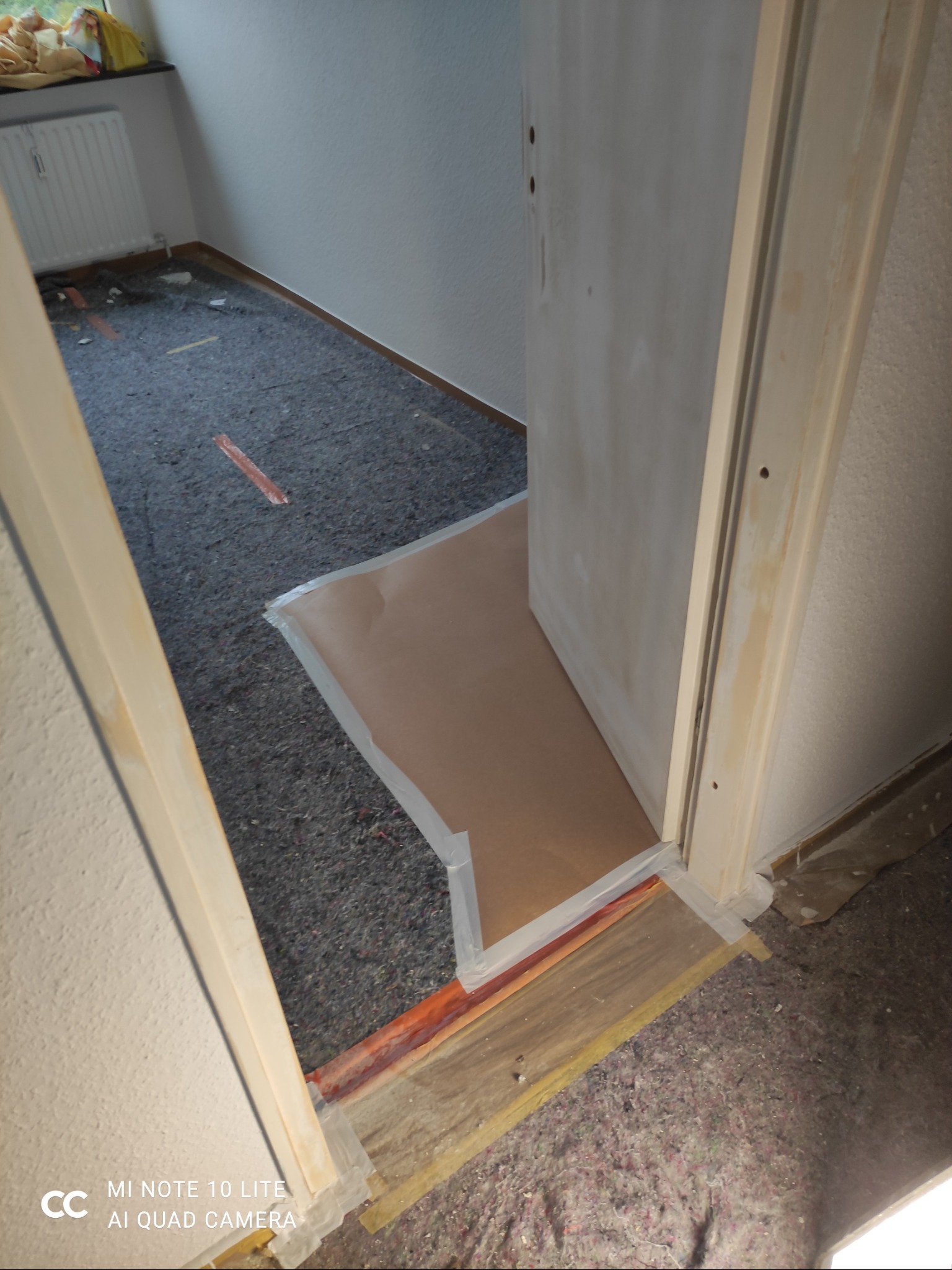 Przygotowanie do malowania: zabezpieczona folią i taśmą podłoga, futryna i dolna część ściany. Pokrycie podłogi szarą włókniną ochronną. Widoczne ślady po szpachlowaniu.