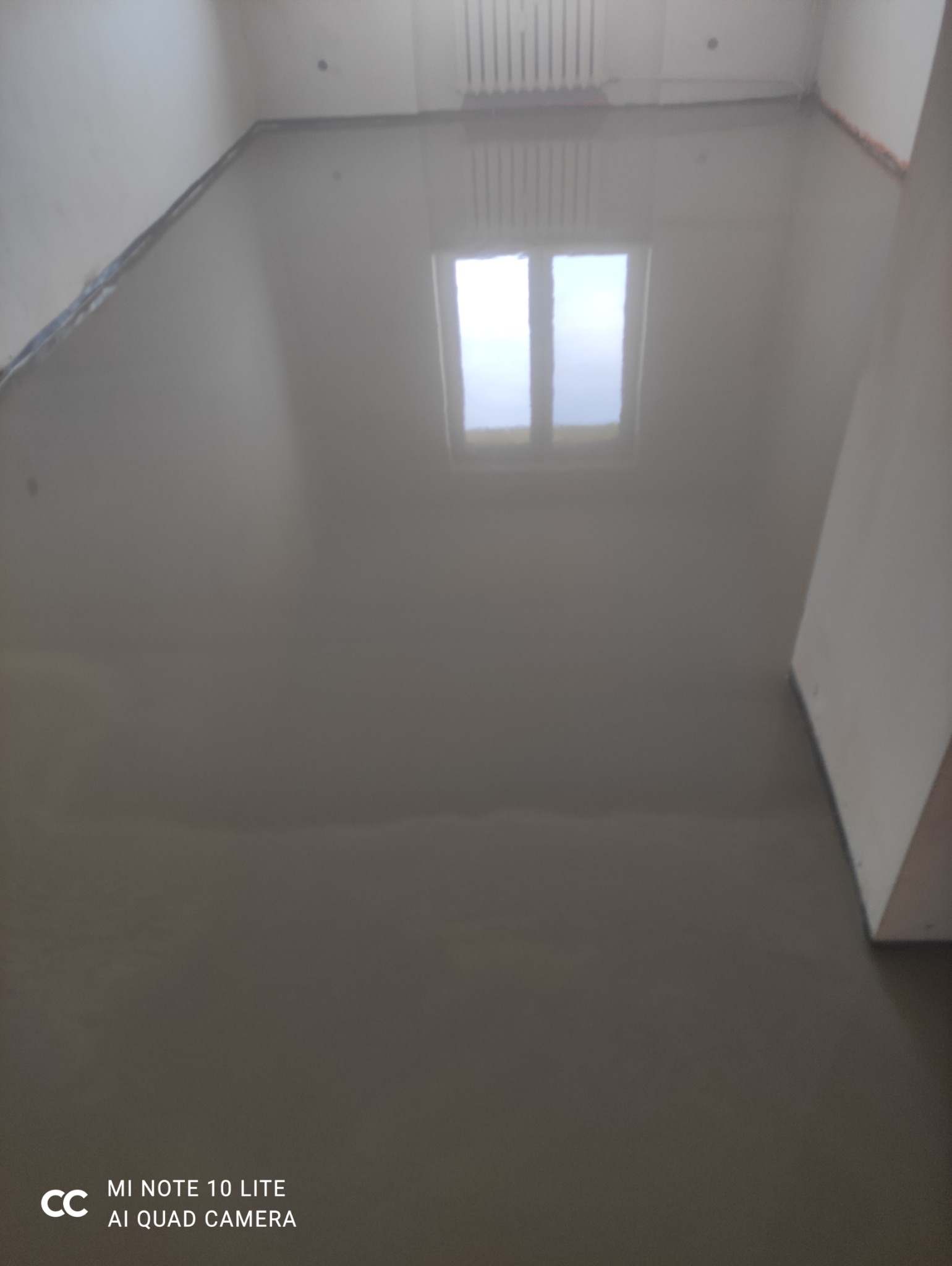 Wylana, gładka posadzka samopoziomująca w pokoju. Widoczne ściany i okno odbijające się w mokrej powierzchni. Na podłodze brak jakichkolwiek zanieczyszczeń.