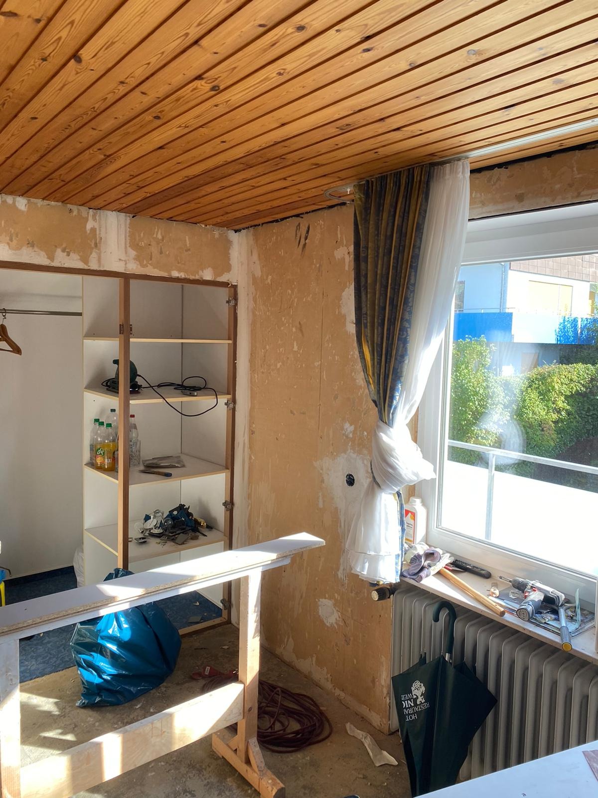 Pokój w trakcie remontu: ściany przygotowane do malowania, widoczne narzędzia, drewniany sufit i częściowo zdemontowana szafa. Widok na zewnątrz przez okno.