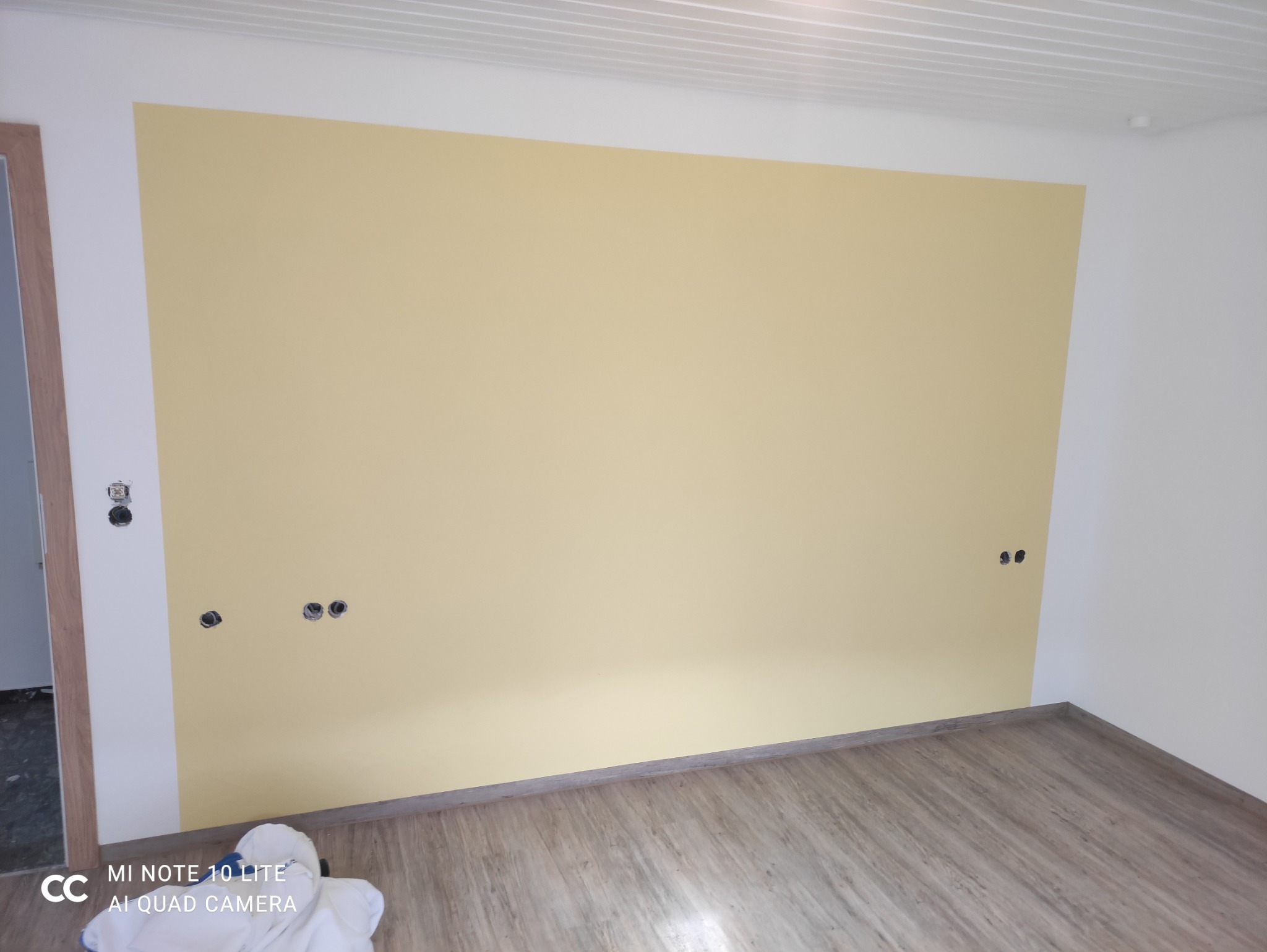 Pomalowana na jasnożółto ściana w pokoju z panelami podłogowymi i gniazdkami elektrycznymi bez osprzętu. Widoczne zabezpieczenia podłogi.