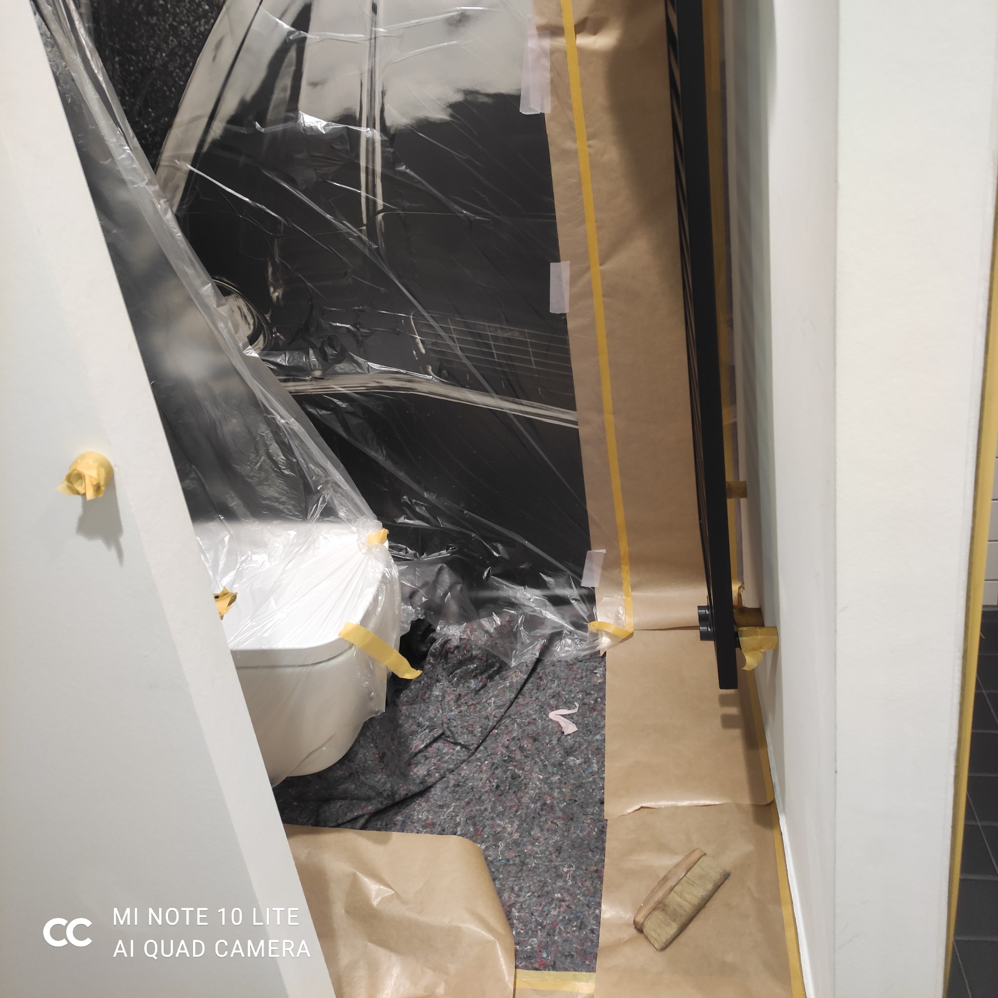 Przygotowanie łazienki do malowania: zabezpieczona folią toaleta, oklejone taśmą malarską futryny i ściany. Widoczny pędzel i podłoga zabezpieczona materiałem.