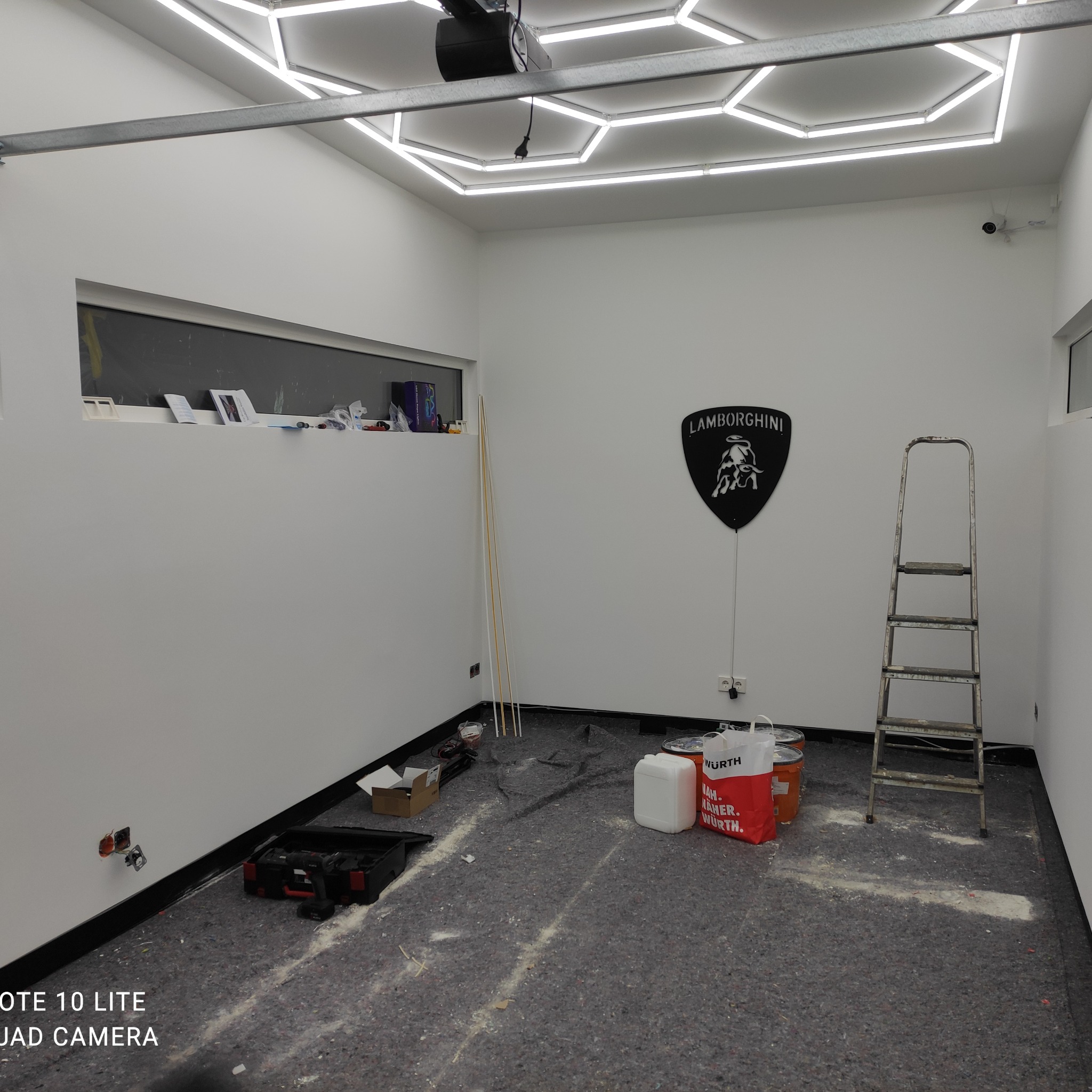 Wnętrze pomieszczenia przed wykończeniem: białe ściany, zabezpieczona podłoga, drabina, narzędzia i logo Lamborghini na ścianie. Widoczne przygotowania do prac.