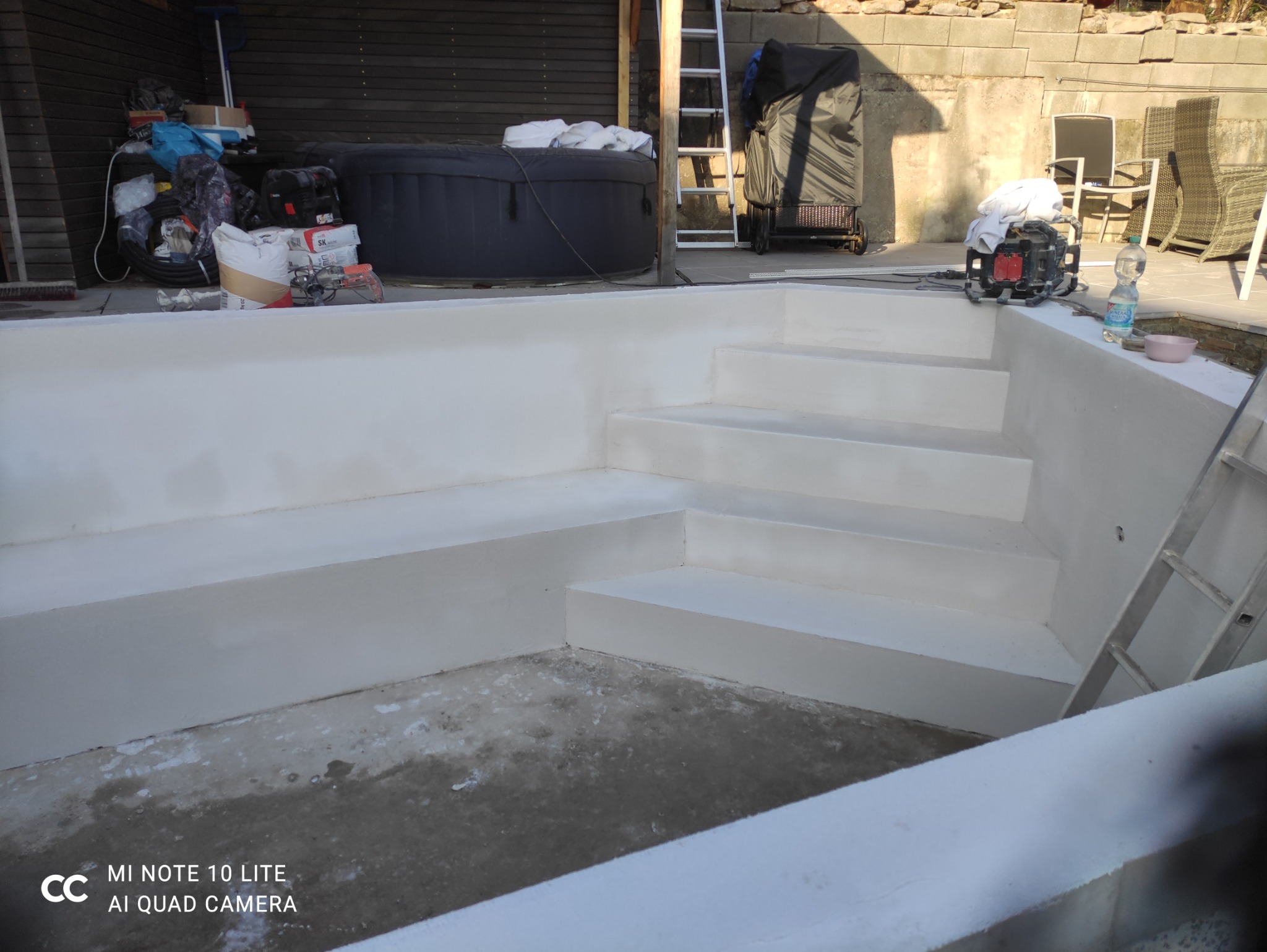 Białe schody basenowe po renowacji, widoczne narzędzia i akcesoria, drabina. W tle jacuzzi i elementy wyposażenia ogrodu. Jasne, naturalne światło.