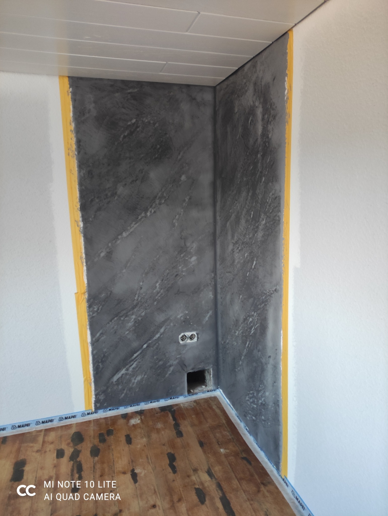 Dekoracyjna szara ściana z efektem marmuru, zabezpieczona taśmą malarską. Widoczne gniazdka elektryczne i fragment drewnianej podłogi. Wykończenie wnętrza.