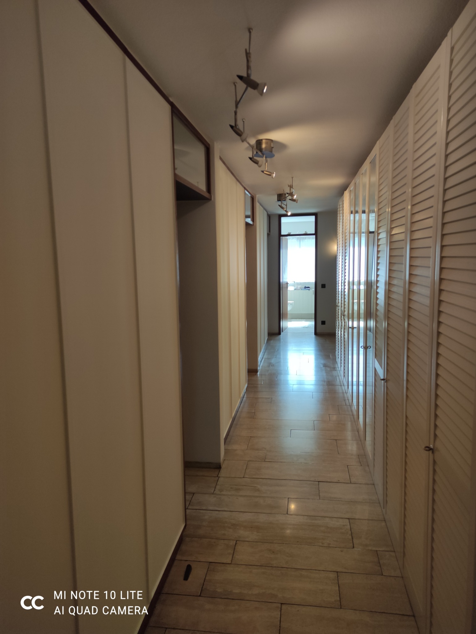 Długi korytarz z zabudowanymi szafami po obu stronach, wyłożony płytkami imitującymi drewno. Oświetlenie punktowe na suficie. Widok na dalsze pomieszczenia.