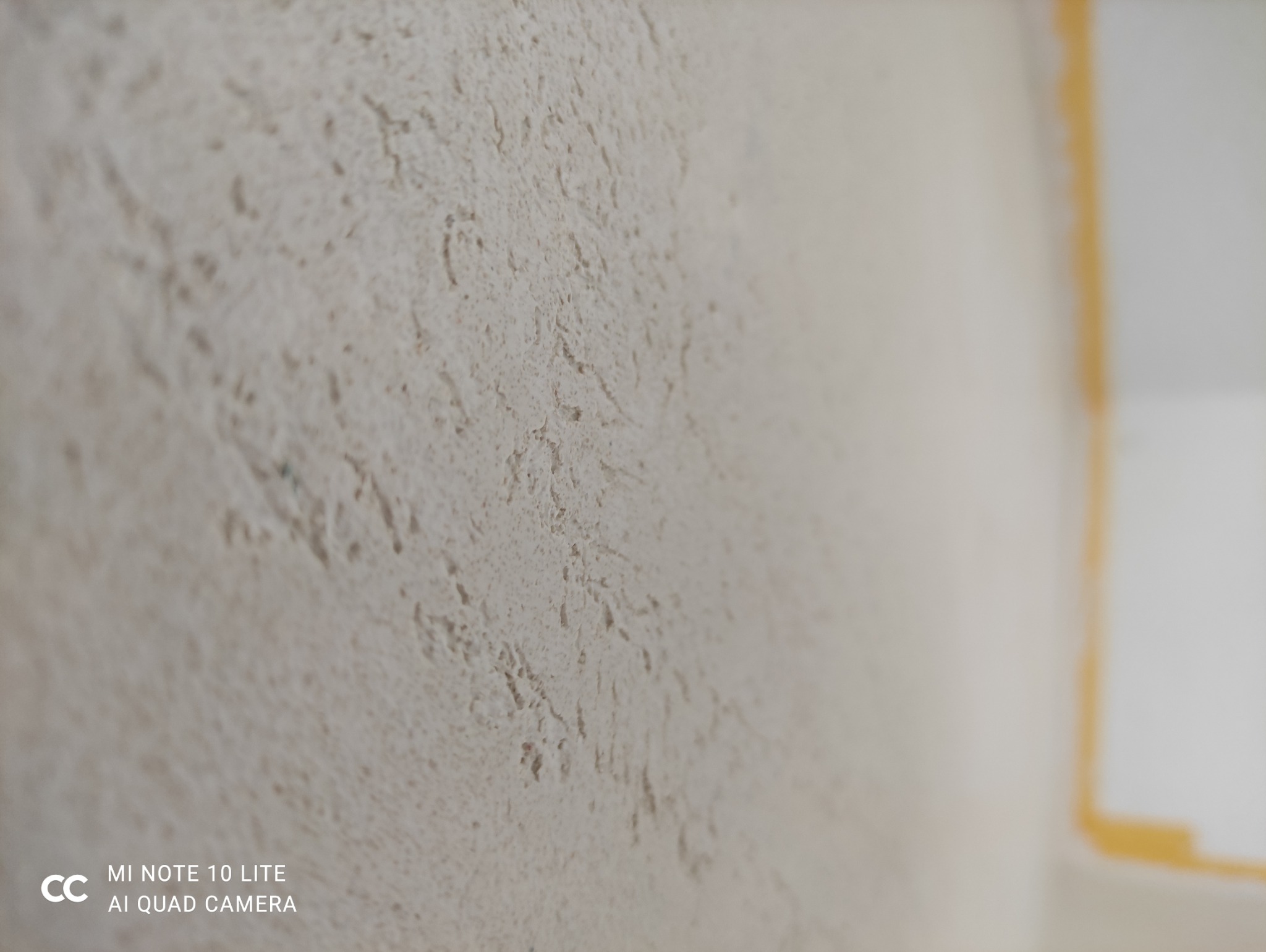 Szpachlowana ściana o nieregularnej fakturze w odcieniach beżu, z widocznym fragmentem żółtej taśmy malarskiej przy suficie. Tekstura ściany jest wyraźnie widoczna.