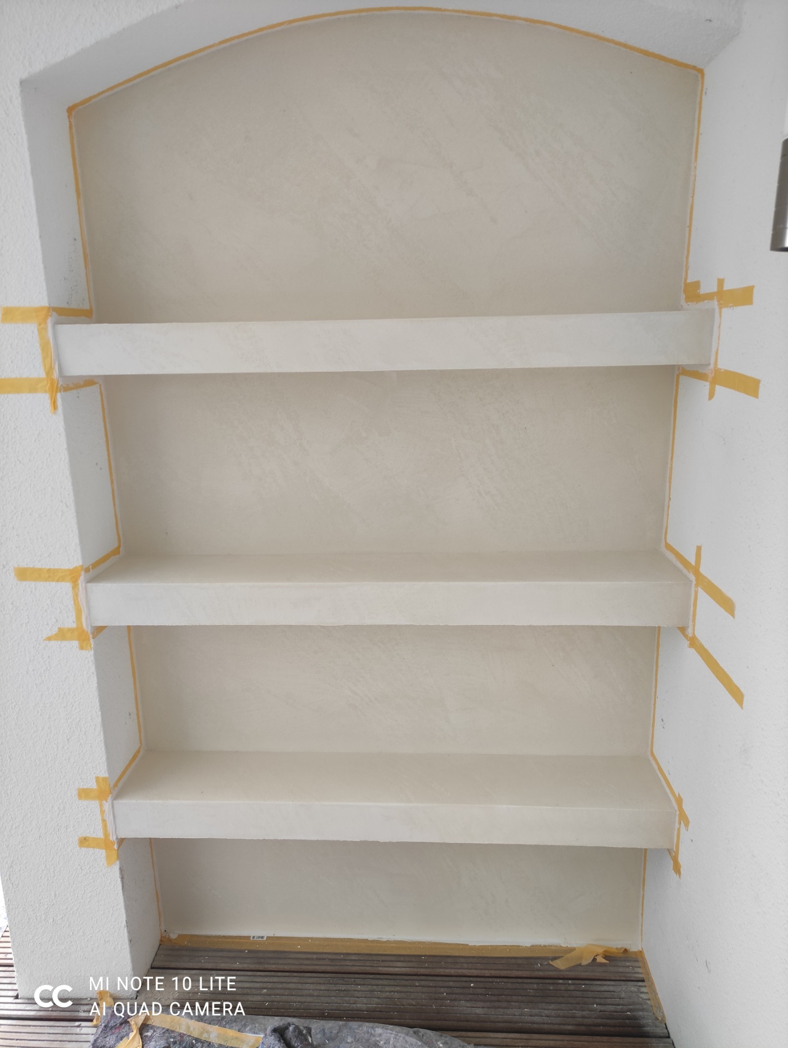 Wnęka z półkami, zabezpieczona taśmą malarską, gotowa do malowania. Widoczne ślady szpachlowania na ścianach i półkach. Przygotowanie do wykończenia wnętrza.