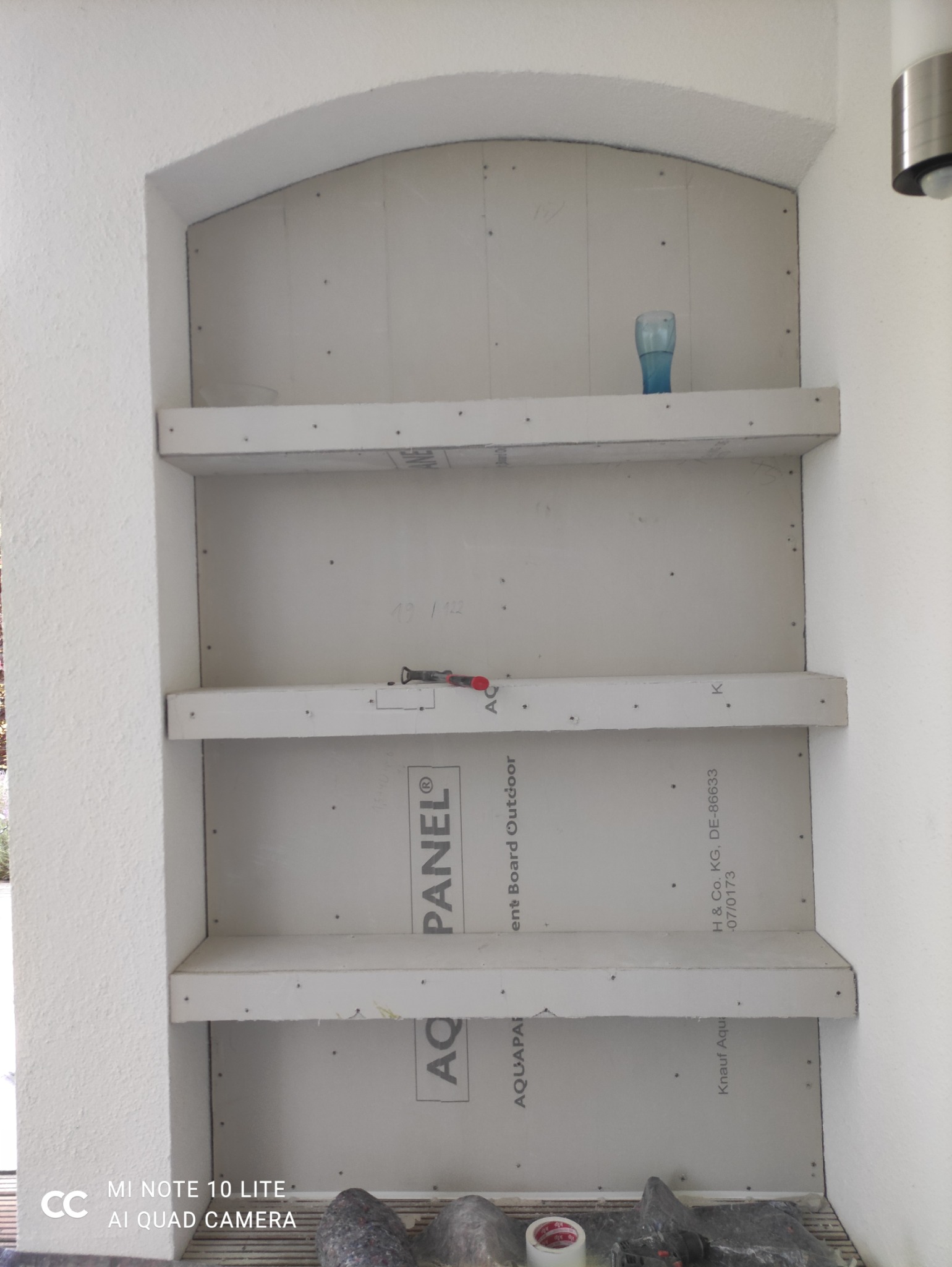 Narożna wnęka z półkami z płyt Aquapanel, w trakcie montażu. Widoczne narzędzia i materiały budowlane. Szara kolorystyka, surowy stan wykończenia.